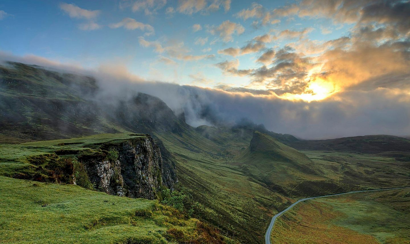 Grassy highland cliffs above a winding road, under low clouds lit by the rising sun