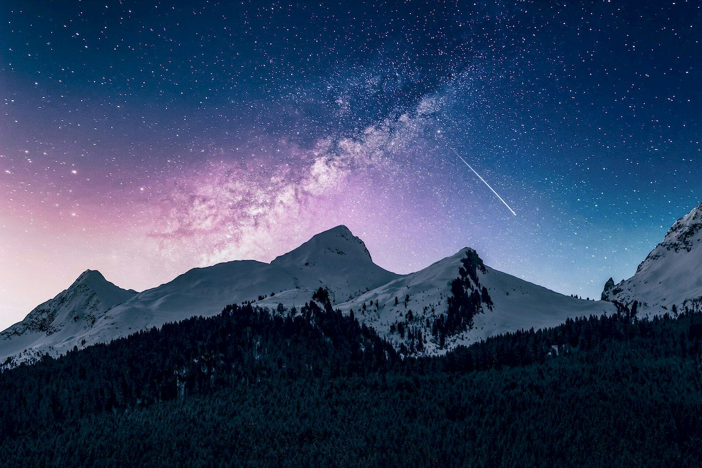 The Milky Way and a shooting star in a night sky over snowy mountains and a dark forest